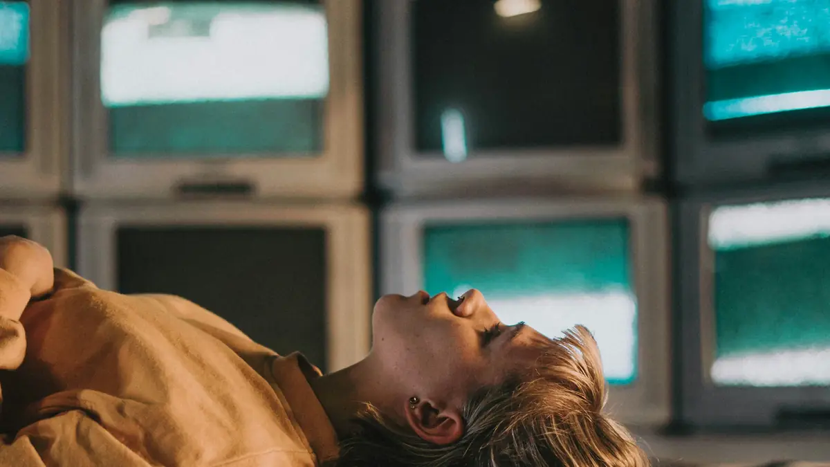A person in a mustard sweatshirt lies back with eyes closed beneath a wall of stacked old CRT televisions glowing with teal static