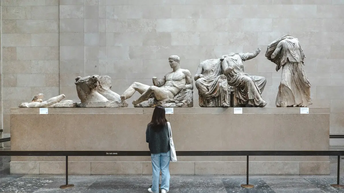 A visitor stands at a rope barrier looking up at the Parthenon sculptures in a stone gallery at the British Museum