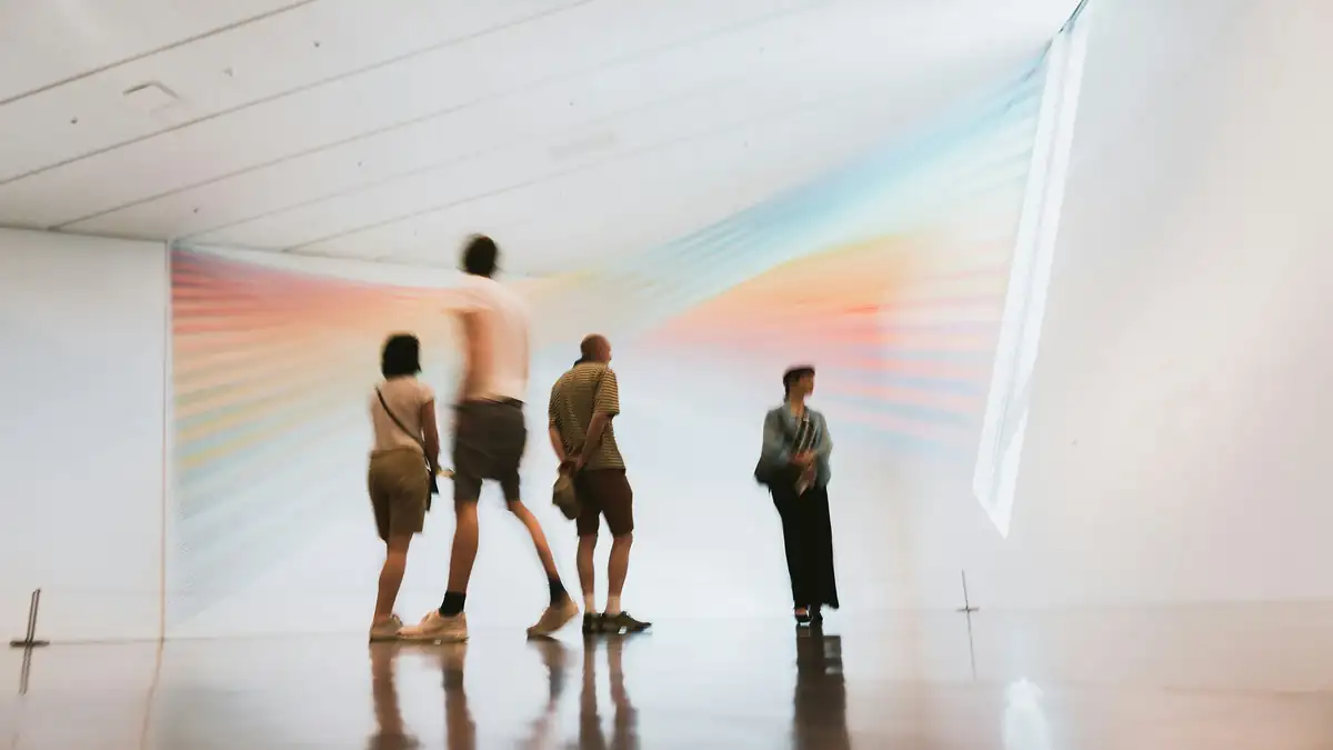 Visitors, blurred in motion, walking through a bright white gallery past a large installation of rainbow-coloured threads